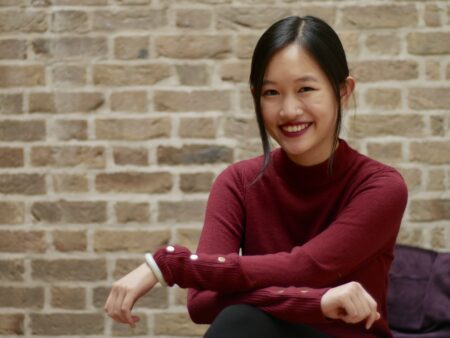 Smiling Asian American woman R.F. Kuang wearing a red shirt while sitting with her arms crossed.